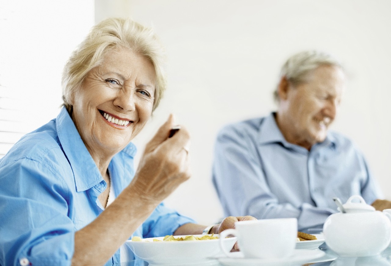 Smiling elderly woman eating a healthy breakfast with a man at the back